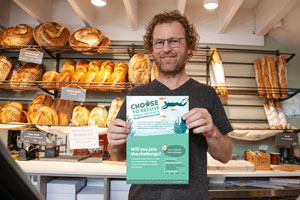 Bakery owner standing in his shop in front of shelves with bread holding up a poster for Plastic Free July reading 'Choose to refuse single use plastics – will you join the challenge?'